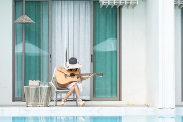 Asian female tourist sits on the edge of her lavish hotel room, playing the guitar and singing,...