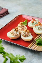 Cold appetizer, eggs stuffed with cream cheese, sun-dried tomatoes, walnuts and herbs on a red ceramic plate on a gray concrete background. Recipes for stuffed eggs.