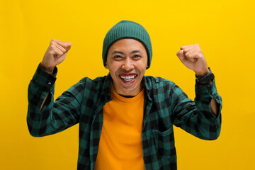 Excited young Asian man, dressed in a beanie hat and casual shirt, is expressing joy and celebrating victory with a fist pump or a YES gesture while standing against a yellow background