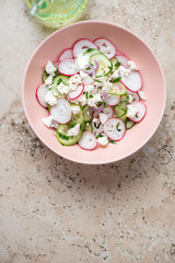 Roseate bowl with cucumber, radish and feta salad, flat lay on a beige granite background, vertical shot, copy space