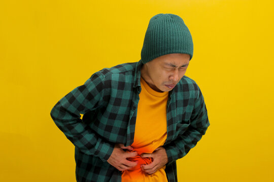 Unhappy Asian man is holding his belly in pain, visibly suffering from a stomachache, likely due to food poisoning, experiencing discomfort, Isolated on yellow background.