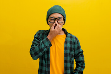 Young Asian man, dressed in a beanie hat and casual shirt, is pinching his nose with his fingers,...