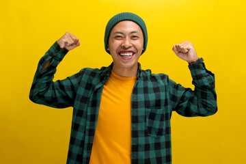 Excited Asian man, dressed in a beanie hat and casual shirt, is making a strong gesture by lifting his arm to show muscles, expressing pride in his achievements, standing against a yellow background