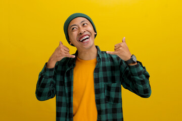 Attractive young Asian man, dressed in a beanie hat and casual shirt, is gesturing a CALL ME sign with his hand while standing against a yellow background