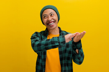 A confident young Asian man, dressed in a beanie hat and a casual shirt, is making a SPEND MONEY gesture while standing against a yellow background