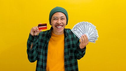 Thrilled Asian man in a beanie and casual clothes holds up a credit card and a stack of banknotes,...