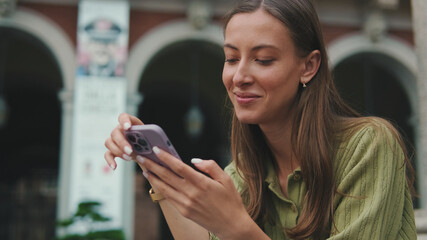 Woman with brown hair, dressed in an olive green sweater, using mobile phone app texting sms on social media