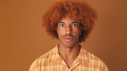 Close up, shocked stylish curly guy asking what while looking at the camera isolated on brown...