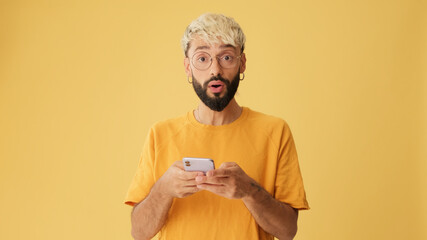 Guy with glasses, dressed in yellow T-shirt, with phone in his hands, surprised, wow effect,...