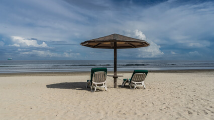 Two deck chairs with soft mattresses stand in the shade of a sun umbrella on a sandy beach. The waves of the turquoise ocean roll towards the shore. Clouds in the blue sky. Malaysia. Borneo.