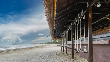 Beach bar by the ocean in the afternoon. Wooden swings are suspended on ropes along the bar counter. Thatched roof on a background of blue sky and clouds. Malaysia. Borneo. Kota Kinabalu