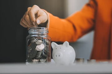 Beautiful Asian businesswoman insert coins in a pin piggy bank on the table. saving money,...