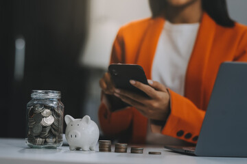 Beautiful Asian businesswoman insert coins in a pin piggy bank on the table. saving money,...