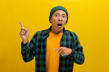 Astonished Asian man, dressed in a beanie hat and casual shirt, points at an empty space, possibly...