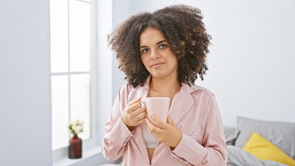 Young hispanic woman with curly hair enjoying coffee in a bright minimalist bedroom.