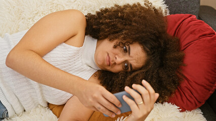 A young hispanic woman with curly hair lounges on a sofa at home, using a smartphone