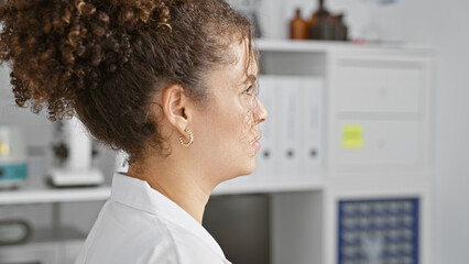 A young hispanic woman with curly hair stands contemplatively in a white lab coat in a clinic setting.