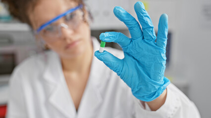 A young hispanic woman with curly hair examines a capsule in a laboratory setting, wearing...