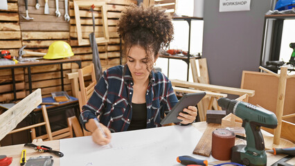 Hispanic woman with curly hair designing in a workshop, surrounded by tools while looking at a...