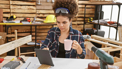 A young hispanic woman with curly hair sips a mug of coffee while studying a digital tablet in a...