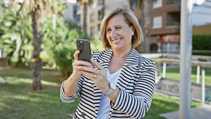 A cheerful mature woman using a smartphone in a green park outdoor setting.