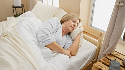 A middle-aged blonde woman looks tired resting in a bright bedroom, evoking themes of health, well-being, and home comfort.