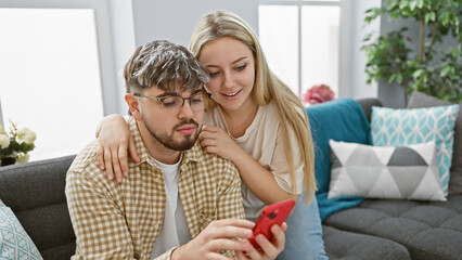 A man and woman cuddle on a sofa in a cozy living room, looking at a smartphone together, radiating...