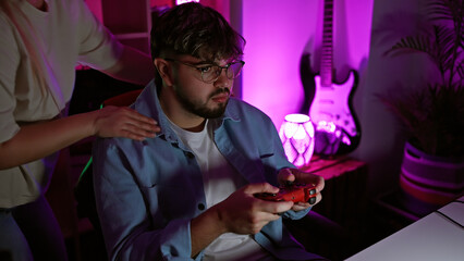 A woman comforts a focused man holding a controller in a dimly lit gaming room at night.