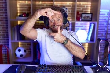 Middle age man with beard playing video games wearing headphones smiling making frame with hands...