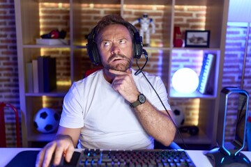 Middle age man with beard playing video games wearing headphones thinking worried about a question,...