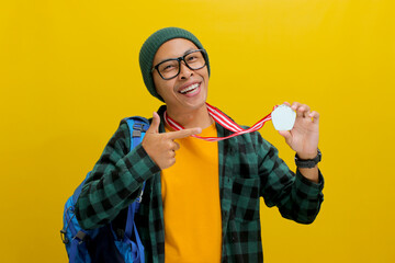 A proud young Asian man, dressed in a beanie hat and casual shirt, smiles as he points at his medal...