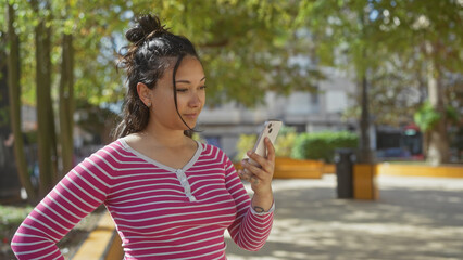 A young latina woman checks her smartphone while standing in a sunny urban park.