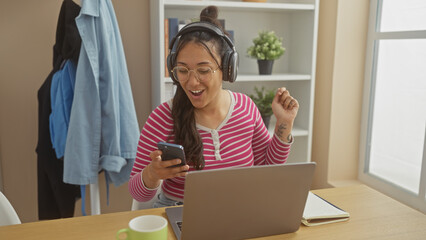 A cheerful young woman listens to music on headphones while using a smartphone and laptop at a home office setting.