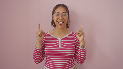 Excited young hispanic woman with glasses pointing up, isolated against a pink wall, wearing a...