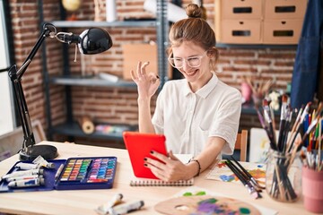 Smiling young blonde artist in studio gestures excellent sign while looking at tablet screen