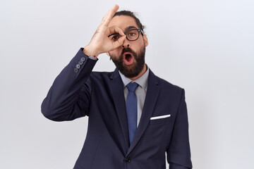 Hispanic man with beard wearing suit and tie doing ok gesture shocked with surprised face, eye looking through fingers. unbelieving expression.