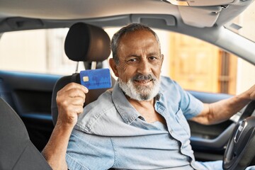 Senior grey-haired man holding credit card sitting on car at street