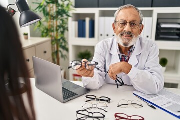 Senior grey-haired man optician holding glasses showing to patient at clinic