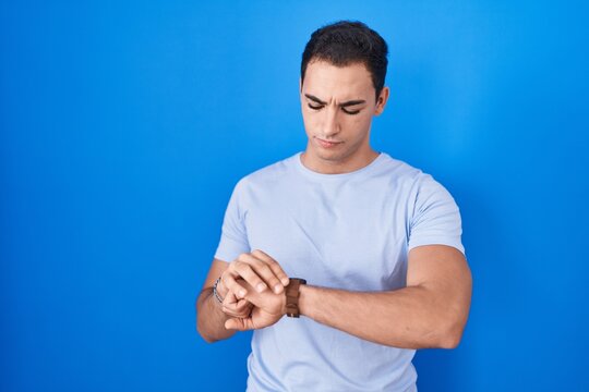 Young hispanic man standing over blue background checking the time on wrist watch, relaxed and confident