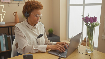 Focused african woman wearing headphones working on laptop at home office with flowers in background