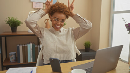 Happy african american woman making bunny ears gesture during a video call in a cozy home office...