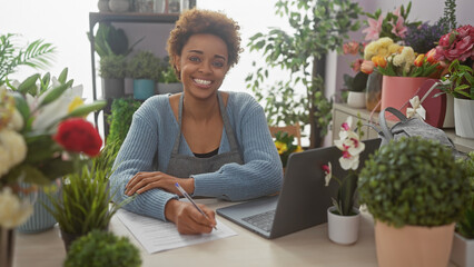 A smiling african american woman entrepreneur writing in a flower shop surrounded by lush plants.