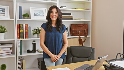 Confident hispanic woman poses in a professional office setting with shelves, laptop, and bookcase.