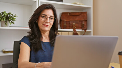 Professional hispanic mature woman working in an office interior with laptop, glasses and elegant attire.