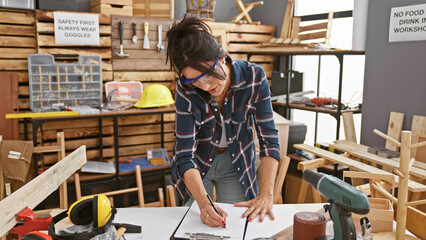 Carpenter woman drawing plans in workshop surrounded by tools and wood.