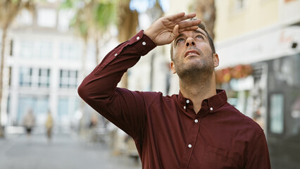 A young hispanic man in a burgundy shirt shields his eyes while standing on an urban city street.