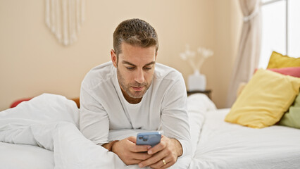 Hispanic man with beard lounging in bedroom using smartphone