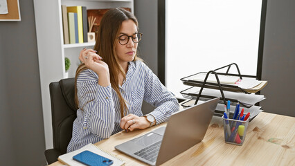 A thoughtful woman with glasses and striped shirt sits at her office desk working on a laptop,...