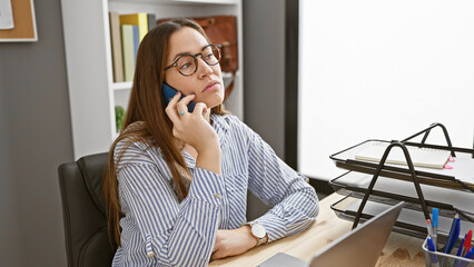 A pensive young woman with glasses on a phone call in a modern office setting, exhibiting a sense...