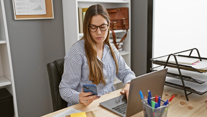 Brunette woman in glasses multitasking with smartphone and laptop in modern office setting